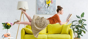 Photo of a woman wearing an orange shirt and tan skirt floating over a yellow couch and reading a paperback book